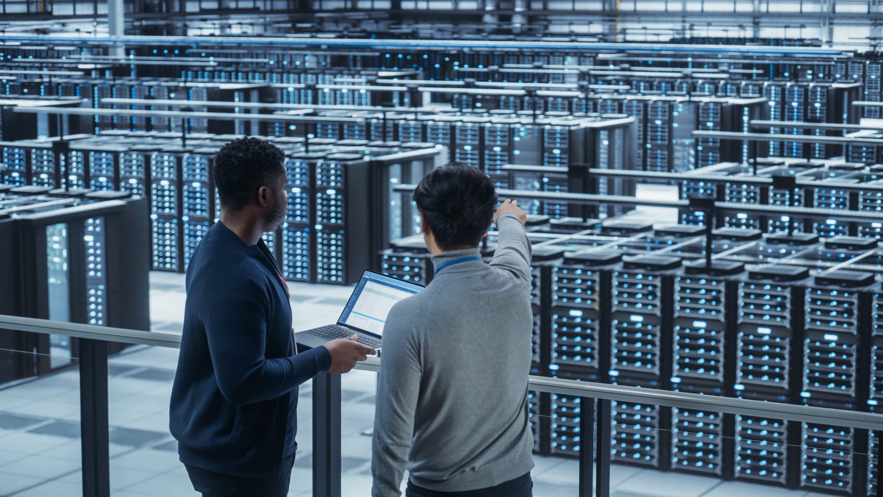 Two engineers with a laptop overlooking a massive hall of blue-lit server racks, one pointing.
