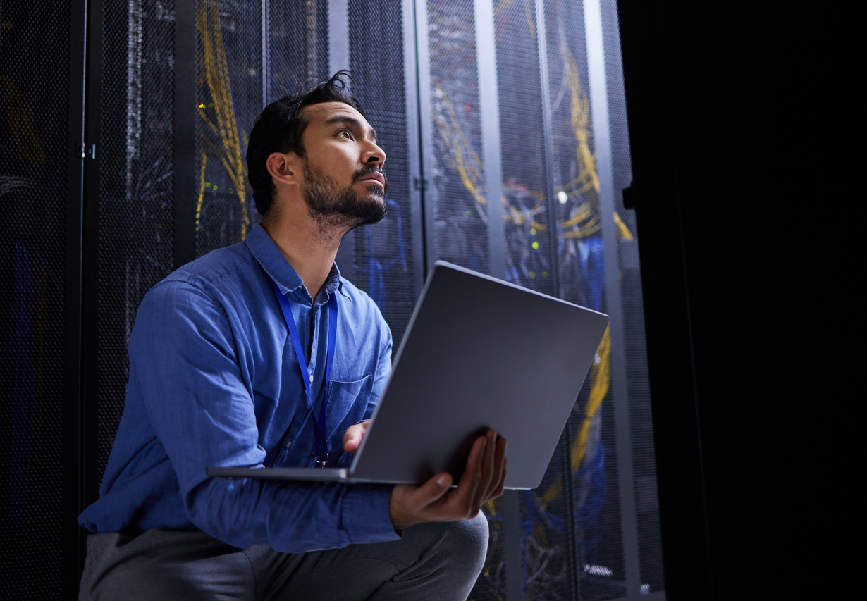 An IT administrator crouched in a dark data center, looking uncertain while holding a laptop amid tangled server cabling
